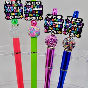 Pode incluir: Quatro canetas coloridas com tampas decorativas e o texto "WE'RE ALL different BUT IN THIS CLASS We swim TOGETHER". As canetas são vermelhas, verdes, roxas e azuis, com glitter e detalhes de contas.