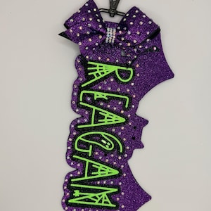 May include: A purple glitter keychain featuring a bow at the top and a black plastic clip. The name REAGAN is written vertically in bright green letters with a spiderweb pattern. The entire surface is covered in small, reflective rhinestones.