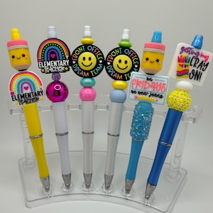 Può includere: Una collezione di penne decorative con vari elementi decorativi, tra cui matite, arcobaleni e faccine sorridenti. Alcune penne presentano il testo "Elementary Teacher", "Front Office Dream Team", "On Fridays we wear jeans", e "Getting my Cray On!"