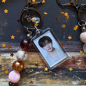 Puede incluir: Llaveros con un dije rectangular transparente con el retrato de una persona. Los llaveros tienen un anillo de metal negro y una cadena de cuentas con cuentas marrones, blancas y rosas. También se adjunta un pequeño dije iridiscente.