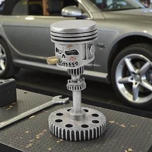 Könnte beinhalten: Eine silberne Metallskulptur eines schädelartigen Kolbens mit einer Kugel im Mund, montiert auf einer zahnradförmigen Basis. Die Skulptur hat eine mechanische Ästhetik mit sichtbaren Motorkomponenten und einem Totenkopf-Gesicht.