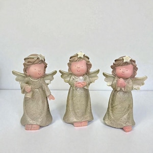 Peut inclure: Trois figurines d'anges avec des robes, des ailes et des cheveux dorés. Chacun a une étoile sur la tête et un visage souriant. L'un tient un parchemin, un autre une colombe, et le troisième a les mains jointes en prière.
