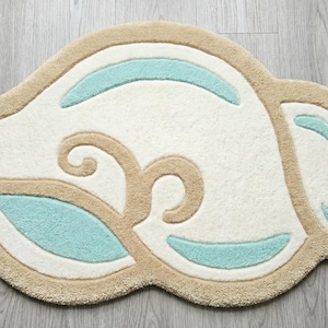 May include: A seashell-shaped rug with a cream-colored center, outlined in tan, and accented with light blue details. The rug is made of a soft, plush material, perfect for a bathroom or bedroom.