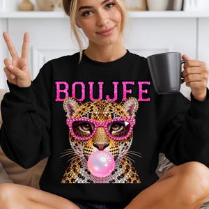 Peut inclure: Un sweat-shirt noir &agrave; col rond avec une impression graphique repr&eacute;sentant un l&eacute;opard portant des lunettes roses scintillantes et faisant une bulle de chewing-gum rose. Le mot BOUJFE est imprim&eacute; en lettres roses scintillantes sur la poitrine.