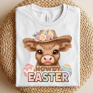 May include: White t-shirt featuring a cartoon cow wearing a straw hat adorned with flowers. The cow is positioned between two Easter eggs, with the words "HOWDY EASTER" printed below in colorful letters. The shirt is folded on a woven surface.