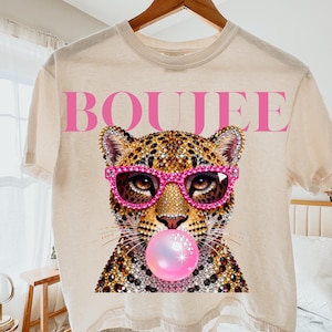 Peut inclure: Un t-shirt couleur cr&egrave;me avec un motif repr&eacute;sentant un l&eacute;opard portant des lunettes de soleil roses orn&eacute;es de strass et faisant une bulle rose. Le mot BOUJEE est imprim&eacute; en grandes lettres capitales roses au-dessus du l&eacute;opard.