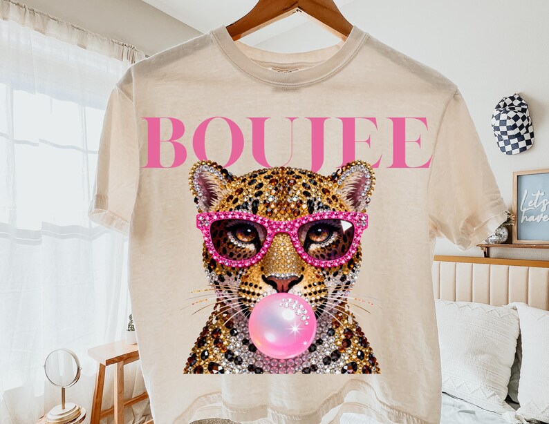 Peut inclure: Un t-shirt couleur cr&egrave;me avec un motif repr&eacute;sentant un l&eacute;opard portant des lunettes de soleil roses orn&eacute;es de strass et faisant une bulle rose. Le mot BOUJEE est imprim&eacute; en grandes lettres capitales roses au-dessus du l&eacute;opard.