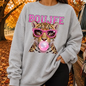 Peut inclure: Un sweat-shirt gris clair &agrave; col rond orn&eacute; d'un motif repr&eacute;sentant un l&eacute;opard portant des lunettes roses &agrave; paillettes et faisant une bulle de chewing-gum rose. Le mot BOUJEE est imprim&eacute; en lettres majuscules roses &agrave; paillettes.
