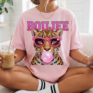 Peut inclure: Un t-shirt rose clair avec un motif repr&eacute;sentant un l&eacute;opard portant des lunettes roses style strass et faisant une bulle de chewing-gum rose. Le mot BOUJFE est imprim&eacute; en grandes lettres majuscules roses et scintillantes au-dessus du l&eacute;opard.
