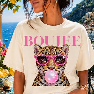 Peut inclure: Un t-shirt &agrave; manches courtes de couleur cr&egrave;me avec le mot BOUJEE en lettres roses au-dessus d'un l&eacute;opard portant des lunettes de soleil incrust&eacute;es de strass roses et faisant une grosse bulle de chewing-gum rose, avec une texture bijou.