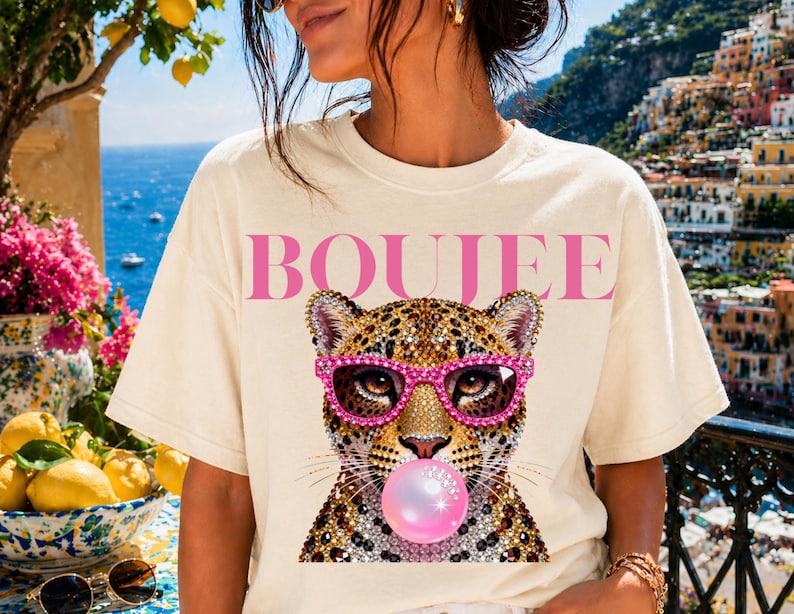 Peut inclure: Un t-shirt &agrave; manches courtes de couleur cr&egrave;me avec le mot BOUJEE en lettres roses au-dessus d'un l&eacute;opard portant des lunettes de soleil incrust&eacute;es de strass roses et faisant une grosse bulle de chewing-gum rose, avec une texture bijou.