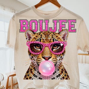 Peut inclure: Un t-shirt couleur cr&egrave;me avec un motif repr&eacute;sentant un l&eacute;opard portant des lunettes rose vif et faisant une bulle de chewing-gum rose. Le mot BOUJFEE est imprim&eacute; en grandes lettres grasses bord&eacute;es de rose au-dessus de la t&ecirc;te du l&eacute;opard. Le design pr&eacute;sente une texture scintillante rappelant des strass sur le pelage et les lunettes.