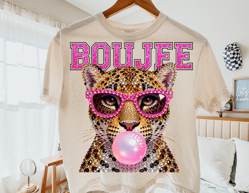 Peut inclure: Un t-shirt couleur cr&egrave;me avec un motif repr&eacute;sentant un l&eacute;opard portant des lunettes rose vif et faisant une bulle de chewing-gum rose. Le mot BOUJFEE est imprim&eacute; en grandes lettres grasses bord&eacute;es de rose au-dessus de la t&ecirc;te du l&eacute;opard. Le design pr&eacute;sente une texture scintillante rappelant des strass sur le pelage et les lunettes.