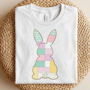 Può includere: T-shirt bianco con un design a coniglietto patchwork. Il coniglietto è composto da vari motivi di tessuto dai colori pastello, tra cui quadretti, floreali e colori uniti. Un piccolo fiocco rosa adorna la testa del coniglietto, creando un look affascinante e festoso.