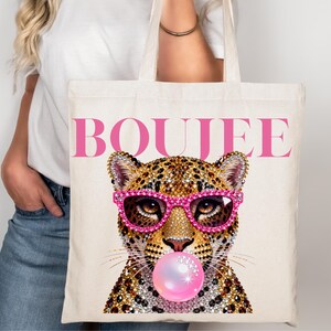 Peut inclure: Un sac fourre-tout en toile couleur cr&egrave;me avec une impression graphique repr&eacute;sentant un l&eacute;opard portant des lunettes incrust&eacute;es de strass rose vif et faisant une bulle de chewing-gum rose. Le mot BOUJEE est imprim&eacute; en grandes lettres roses.