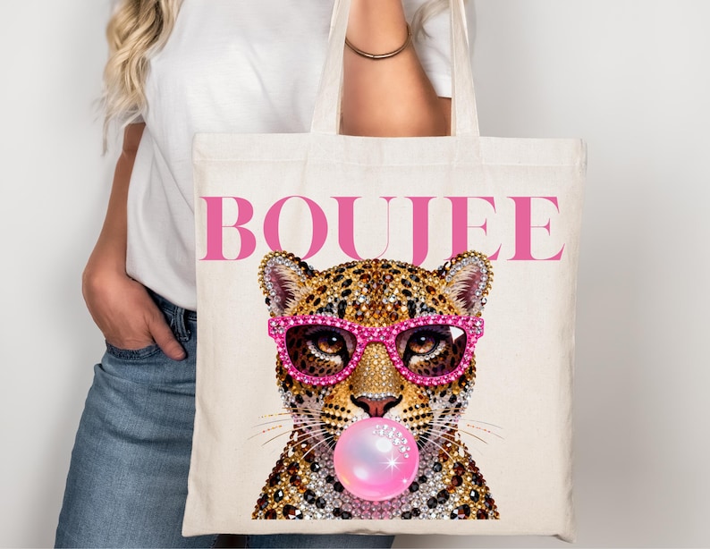 Peut inclure: Un sac fourre-tout en toile couleur cr&egrave;me avec une impression graphique repr&eacute;sentant un l&eacute;opard portant des lunettes incrust&eacute;es de strass rose vif et faisant une bulle de chewing-gum rose. Le mot BOUJEE est imprim&eacute; en grandes lettres roses.