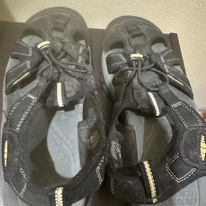 May include: Pair of black and gray outdoor sandals with adjustable straps and a bungee cord closure. The sandals have a textured sole and are designed for water activities. The straps have white and yellow accents.