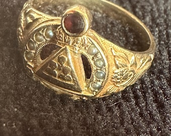 May include: A gold ring with detailed patterns and a central dark red gemstone. The ring has small, light-coloured stones and floral and geometric designs. The overall style suggests a vintage or antique piece, with intricate craftsmanship.