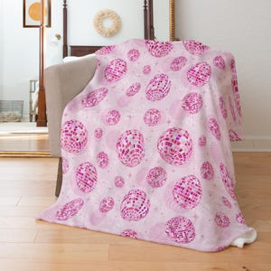 May include: A pink blanket featuring a pattern of disco balls in shades of pink and white. The design includes stars and the name "KYLAN" in a repeating pattern. The blanket is draped over a chair.