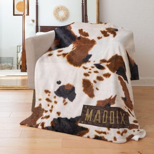May include: A plush blanket with a cowhide pattern in white, brown, and black. The blanket is draped over a chair and features a rectangular patch with the name "MADDIX" in gold lettering.
