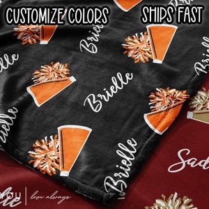 May include: A black blanket with a repeating pattern of orange and white cheerleading megaphones and pom-poms. The name "Brielle" is written in white script across the blanket. The words "Customize Colors" and "Ships Fast" are also visible.