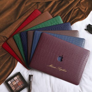May include: A collection of colorful laptop cases in red, brown, green, navy, and burgundy, arranged on a white surface. The cases have a textured, crocodile-like pattern. A pair of black sunglasses and makeup are also present.