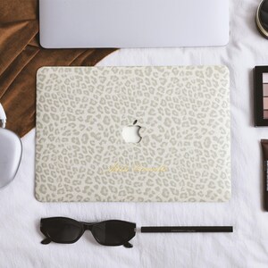 May include: A laptop skin featuring a light beige and white leopard print pattern with a central Apple logo cutout. The skin displays the cursive text "Miss World" in gold lettering. The laptop is surrounded by headphones, sunglasses, and makeup.