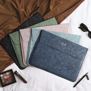 May include: A collection of five rectangular laptop sleeves in black, sage green, dusty pink, light blue, and navy blue, each featuring an embossed floral pattern. The navy blue sleeve is personalized with the gold-colored initials D.B.