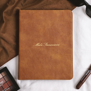 May include: A rectangular, tan-coloured, textured notebook cover featuring the name Mila Genevieve embossed in gold script lettering on the front centre. The cover has rounded corners and a smooth, consistent finish across its surface.