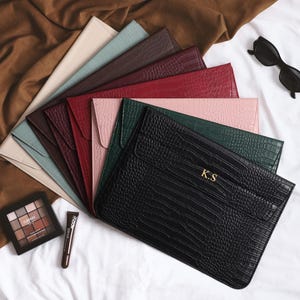 May include: A collection of leather laptop sleeves in various colors, including black, green, red, and pink. The black sleeve features gold initials. Sunglasses, eyeshadow, and a tube of makeup are also present.