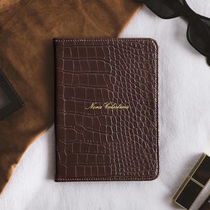 May include: A dark brown rectangular notebook with a textured crocodile skin pattern and the name Nora Celestina embossed in gold lettering on the front cover. The notebook features stitched edges and a glossy finish, presented in a vertical orientation.