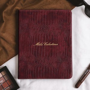 May include: A rectangular, deep burgundy notebook with a textured crocodile-skin pattern and the gold-embossed name Mila Celestina centred on the front cover. The notebook is positioned alongside a black eyeshadow palette and a brown lip treatment tube.