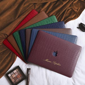 May include: A collection of laptop cases featuring a textured crocodile skin pattern in various colors including red, brown, green, blue, and burgundy. The burgundy case is personalized with the gold text Marie Sophia.