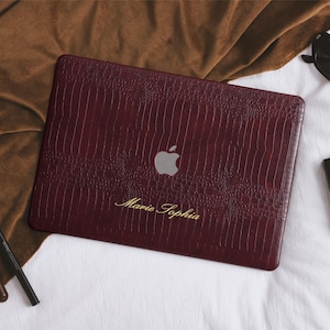 Może przedstawiać: Bordowa obudowa na laptopa o fakturze skóry krokodyla, z logo Apple i imieniem "Marie Sophia" w złotym skrypcie. Obudowa leży na białej powierzchni, w pobliżu okulary przeciwsłoneczne i makijaż.
