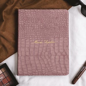 Puede incluir: Una funda para cuaderno rectangular de color rosa empolvado con un patrón texturizado de piel de caimán. El nombre Marie Sophia está grabado en letras doradas cursivas en el centro de la portada. El artículo tiene bordes cosidos.