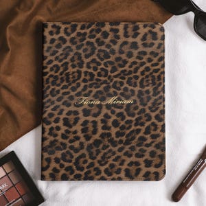 May include: A rectangular notebook cover featuring a brown and black leopard print pattern. The name Fiona Miriam is embossed in gold lettering in the center of the cover. The notebook is positioned alongside a makeup palette and a lip treatment.