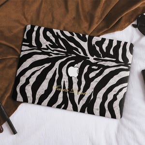 Puede incluir: Una funda para portátil con un patrón de rayas de cebra en blanco y negro, un recorte central para el logotipo de Apple y el texto cursivo dorado Carla Dante impreso cerca del borde inferior de la cubierta protectora rectangular.