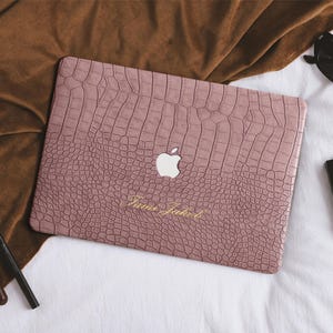 Può includere: Una custodia rosa per laptop con una texture a pelle di coccodrillo, con il logo Apple e il nome "Jamie Jakob" in oro. La custodia è su una superficie bianca, con occhiali da sole e trucco.
