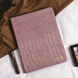 Puede incluir: Un cuaderno de color rosa empolvado con un patrón texturizado de piel de cocodrilo y las iniciales L.B. grabadas en oro en la esquina inferior derecha. El cuaderno es rectangular con esquinas redondeadas y un acabado liso y uniforme.
