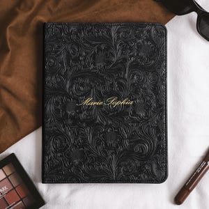 Puede incluir: Un cuaderno rectangular negro con un patrón floral en relieve en la cubierta y el nombre Marie Sophia impreso en letra dorada en el centro. El cuaderno tiene esquinas redondeadas y una superficie texturizada con detalles de hojas en espiral.
