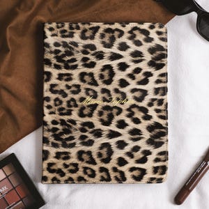Puede incluir: Un cuaderno rectangular con un estampado de leopardo en tonos beige y negro, que presenta el nombre Marie-Sophie grabado en letras doradas cursivas en el centro de la portada, en orientación vertical con esquinas redondeadas.