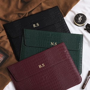 Pode incluir: Três capas para laptop em couro sintético preto, verde e bordô, cada uma em relevo com um padrão de crocodilo. As capas são personalizadas com iniciais douradas. Maquiagem e óculos de sol também são visíveis.