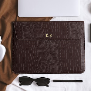 Könnte beinhalten: Eine dunkelbraune Laptoptasche mit Krokodiltextur und den goldenen Initialen "K.S." steht im Mittelpunkt. Das Bild zeigt auch einen weißen Laptop, eine schwarze Sonnenbrille, eine Make-up-Palette und ein weißes Kopfhörerpaar. Die Artikel sind auf einer weißen Oberfläche angeordnet.