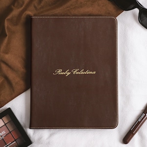 May include: A rectangular dark brown leather portfolio with rounded corners and visible stitching along the edges. The name Ruby Celestina is embossed in gold script lettering in the centre of the front cover of the portfolio.