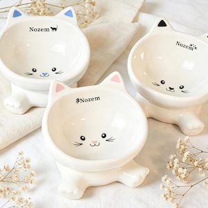 May include: Three white ceramic cat-shaped pet bowls. Each bowl has a cat face design with whiskers and the word "Nozem" printed on the front. The ears are accented with blue, pink, and black. The bowls are on a white surface.