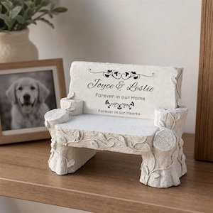 May include: A white stone-look bench ornament featuring a textured tree stump design with vines. The backrest is inscribed with the text Joyce & Leslie, Forever in our Home, and Forever in our Hearts, accented by decorative floral flourishes.