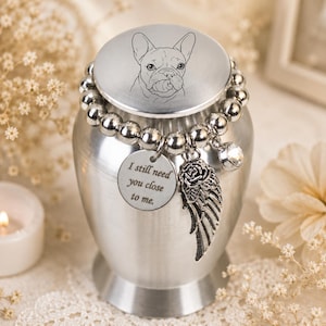May include: A silver-toned metal urn featuring a lid engraved with a French Bulldog illustration. A beaded silver bracelet wraps around the neck, adorned with a round charm inscribed with "I still need you close to me" and a wing charm.