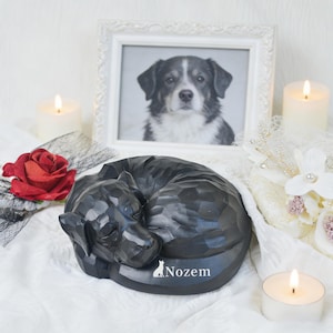 May include: A black, carved dog sculpture in a curled sleeping position, with the word "Nozem" and a dog silhouette on the base. A framed black and white photo of a dog is in the background, along with candles and a red rose.
