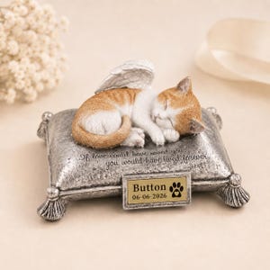 Pode incluir: Uma estatueta de gato laranja e branco a dormir com asas de anjo sobre uma almofada prateada texturizada. A almofada apresenta o texto gravado If love could have saved you, you would have lived forever e uma placa com Button 06-06-2026.