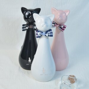 以下が含まれることがあります： 黒、白、ピンクの3つのセラミック製の猫の置物。各猫は、真珠のアクセントが付いた蝶ネクタイをしています。白い猫は青いチェック柄の蝶ネクタイをしています。黒い猫は白と黒のチェック柄の蝶ネクタイをしています。ピンクの猫はピンクと黒のチェック柄の蝶ネクタイをしています。